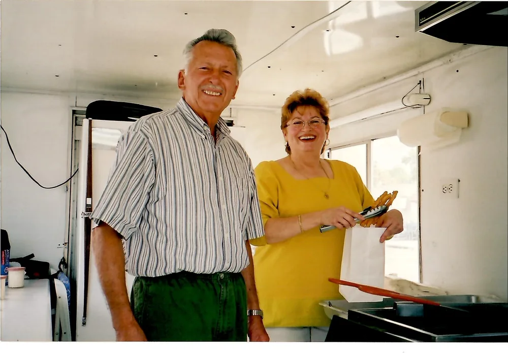 Jacinto y Mari Ramos sonriendo dentro del primer food truck de Churroman en 2001, mientras Mari empaca churros frescos con unas pinzas