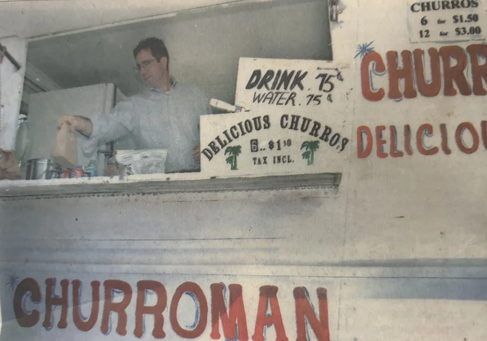 Foto de periódico de 2006 de Eliu Castillo atendiendo en la ventanilla de Churroman bajo letreros pintados a mano: Delicious churros, 6 por $1.50, impuesto incluido